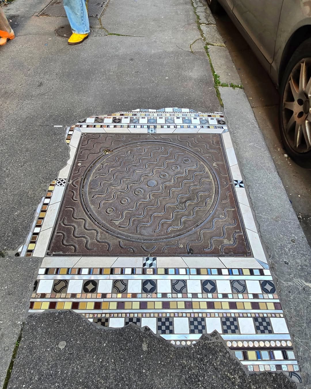 Mosaic surrounding a large circular manhole with linear tile patterns in white, gold, and black, embedded in a cracked urban sidewalk.