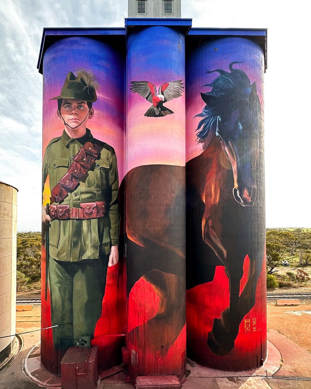 Silo art by Julian Clavijo and Camilo Delgado in Walpeup, Australia, depicting a young WWI soldier and a horse under a vivid sunset with a bird above, all painted across three grain silos.