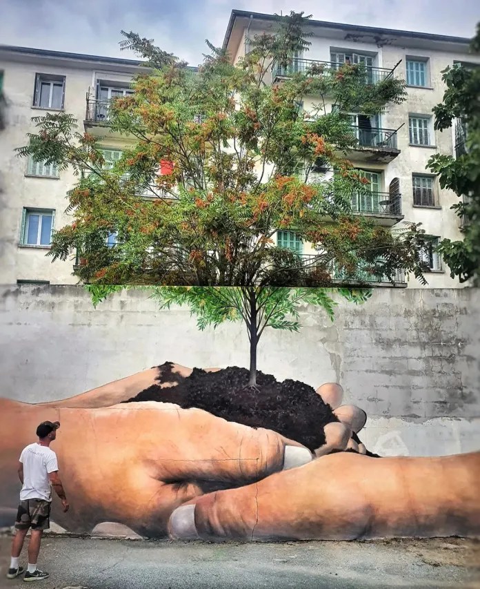 A painted mural of two open hands holding the green leaves of a real tree planted in front of the wall in Ajaccio, France.