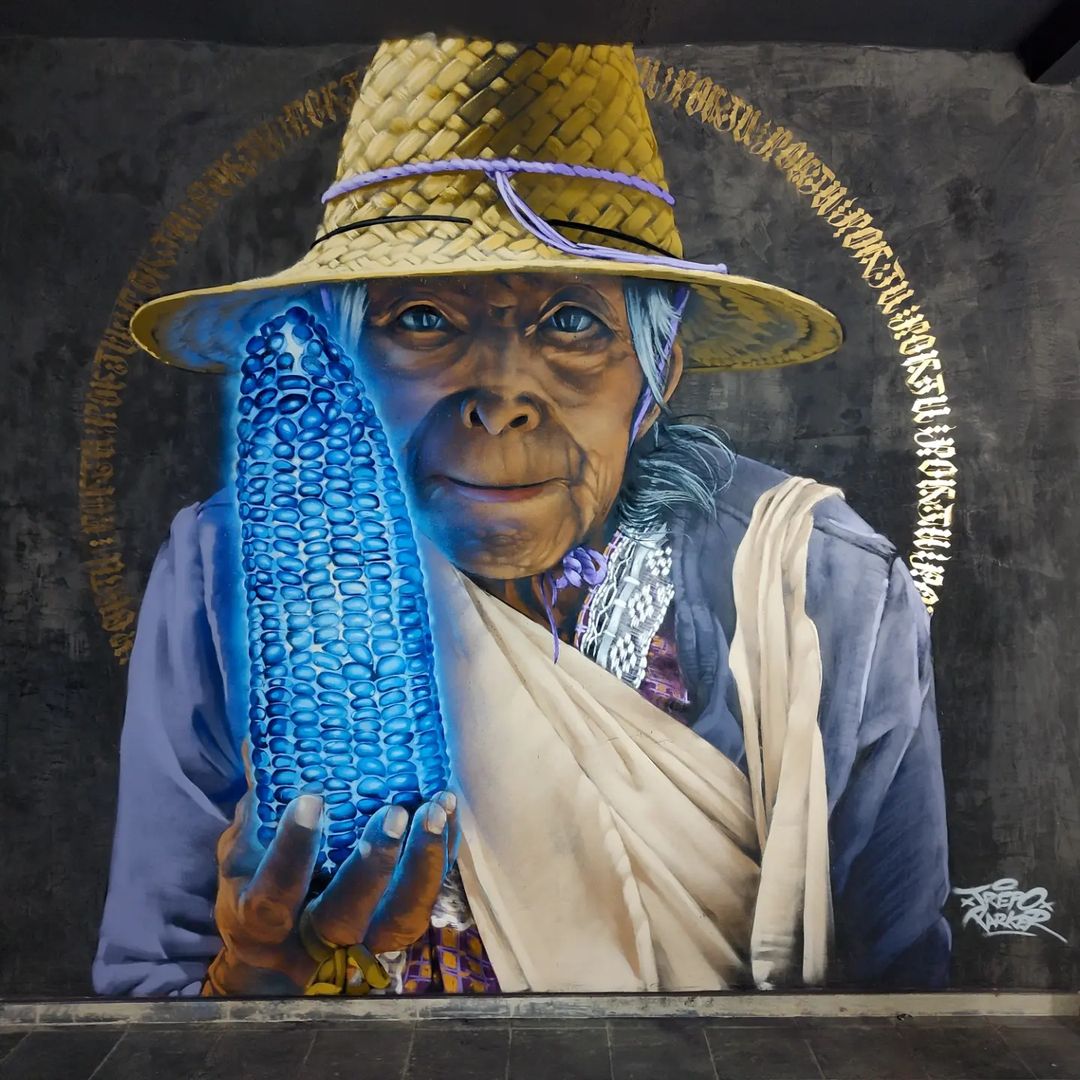 Mural of an indigenous woman holding a glowing blue corn cob, wearing a straw hat and traditional garments, with a golden halo-like arc of script surrounding her head.