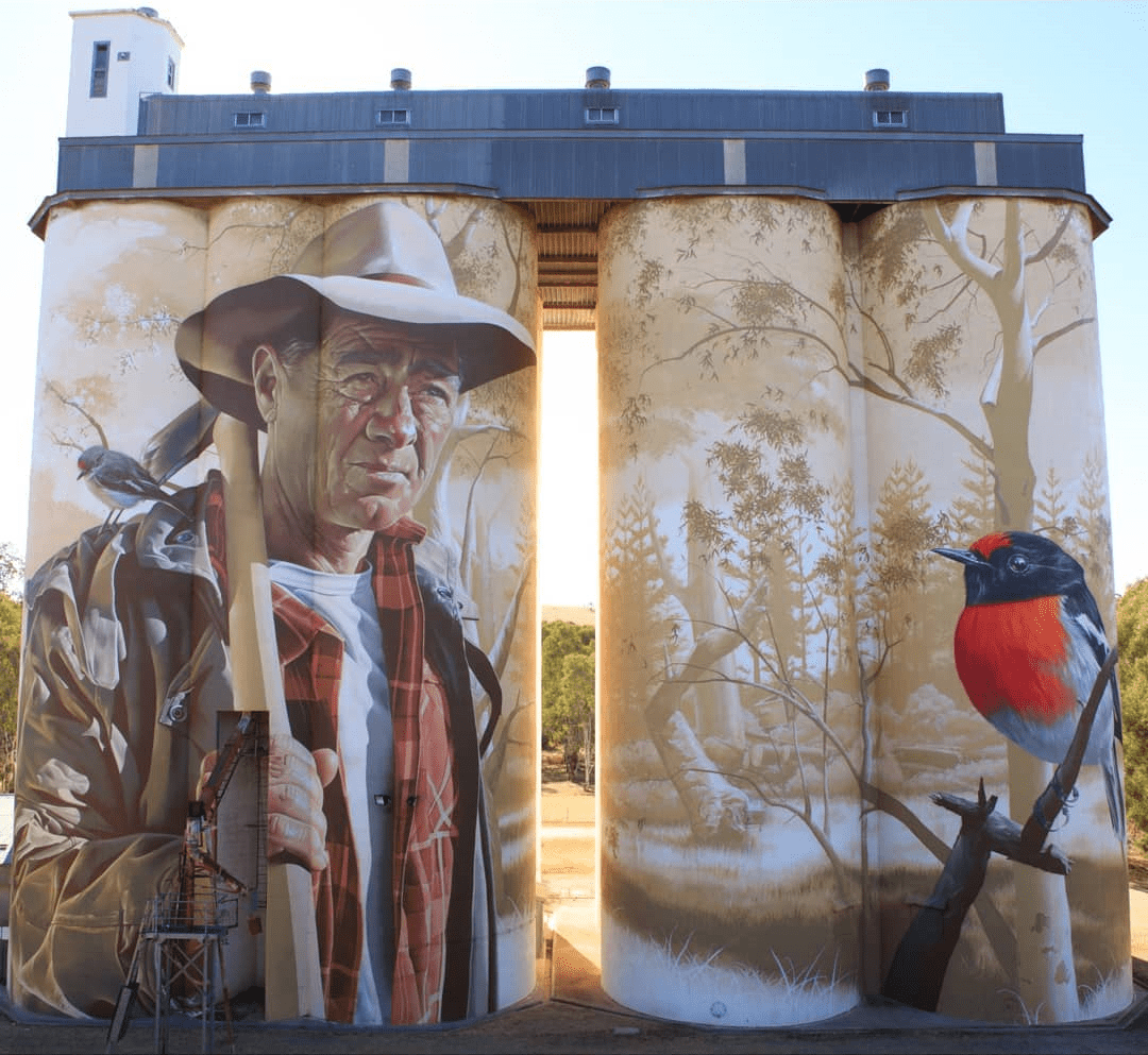 Giant silo mural of a male farmer holding a shovel, with realistic details of his face and clothes. A robin bird appears on a nearby silo, surrounded by faint tree illustrations.