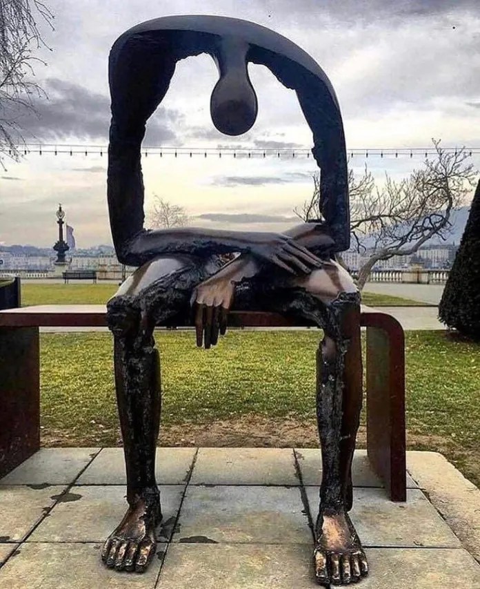 Bronze sculpture of a seated person on a bench in Geneva, Switzerland, by Albert György. The figure has a large hollow void through its torso and head, symbolizing internal emptiness. Background shows a park and lake with cloudy sky.
