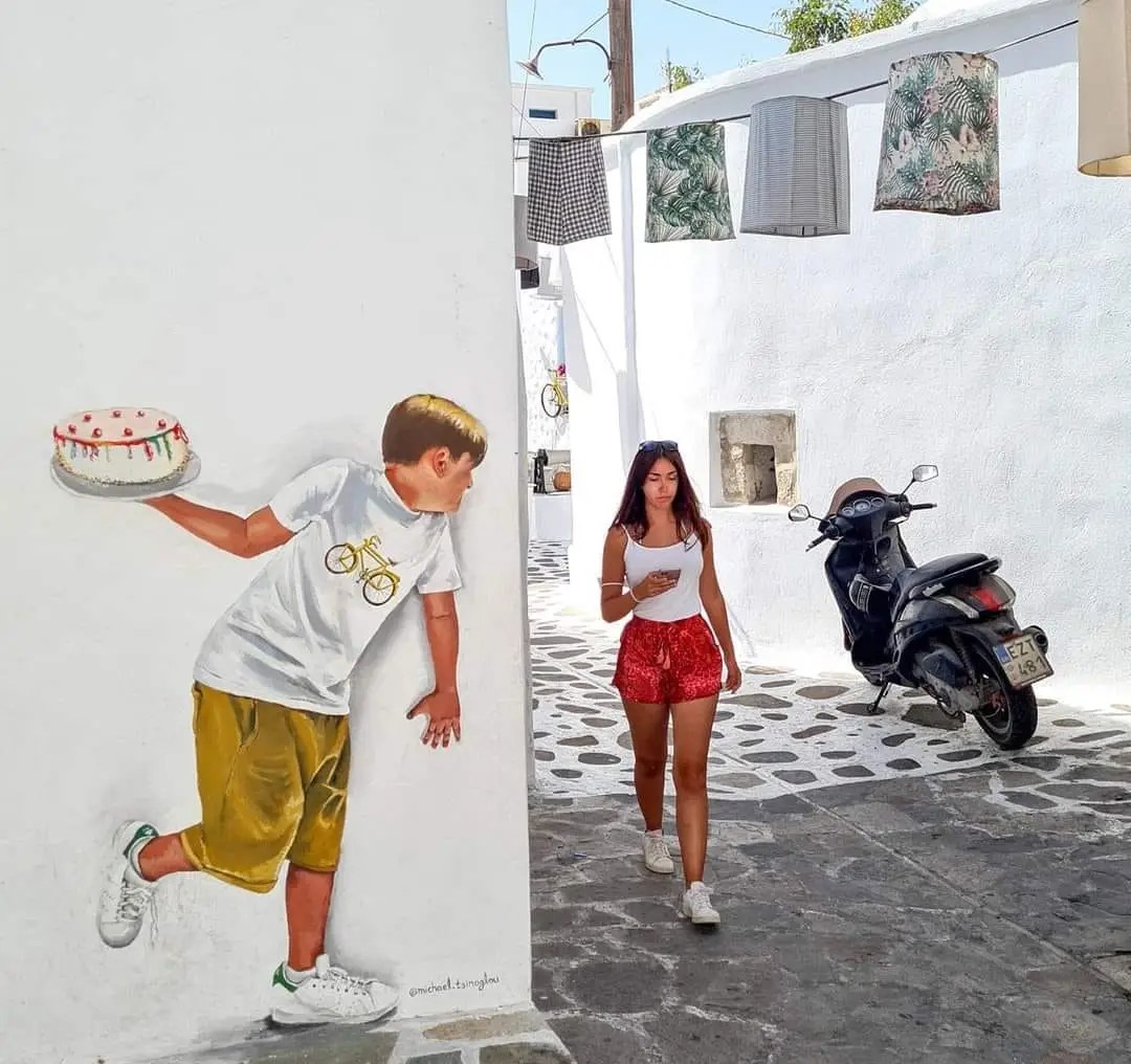 Street art mural in Naxos, Greece by Michael Tsinoglou showing a boy with blond hair holding a cake and peeking around a white corner wall, perfectly positioned to surprise a woman walking down the alley.