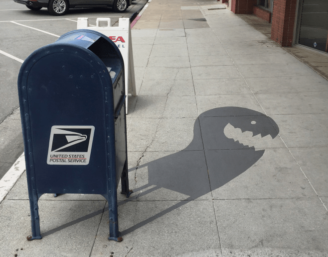 Blue United States Postal Service mailbox on a sidewalk, with a painted shadow extending onto the pavement, shaped like an open-mouthed monster with sharp teeth.