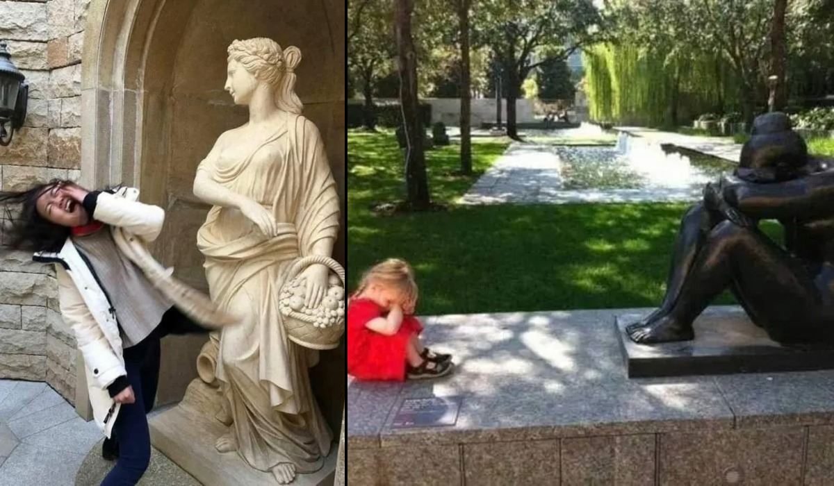 A woman playfully mimics the pose of a stone statue of a woman holding a basket in a garden setting, with trees and a pathway visible in the background.