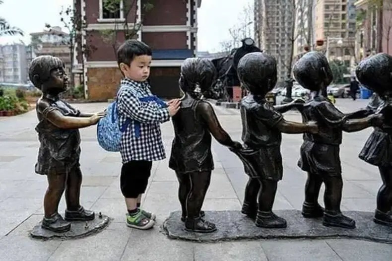 A real child with a blue backpack stands between two bronze children statues holding hands in a queue formation.