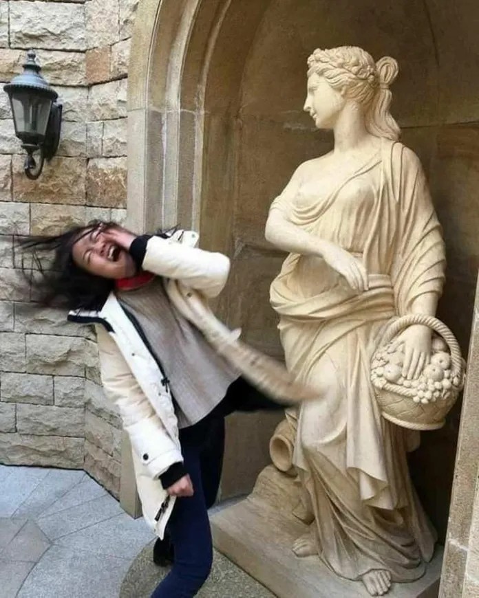 A woman recoils dramatically in front of a classical stone statue holding a fruit basket.