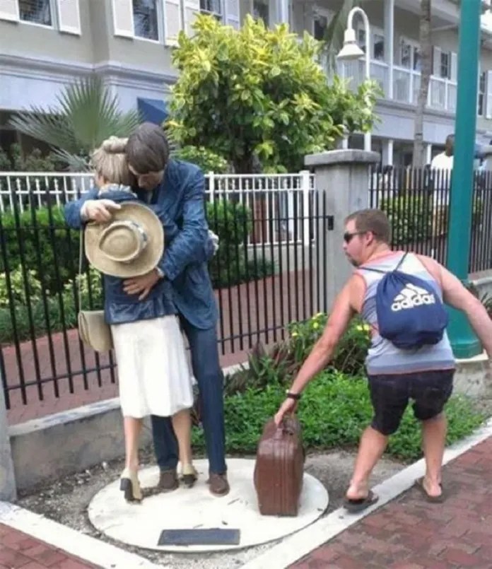 A man leans to grab a suitcase from a sculpture of a hugging couple.