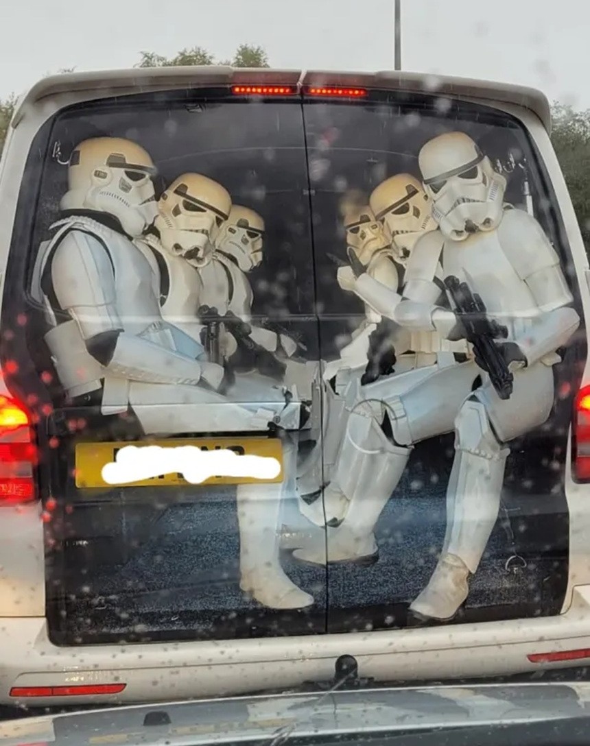 Rear window of a van covered with an image of several Star Wars stormtroopers sitting inside and holding blasters, seen through a wet windshield.