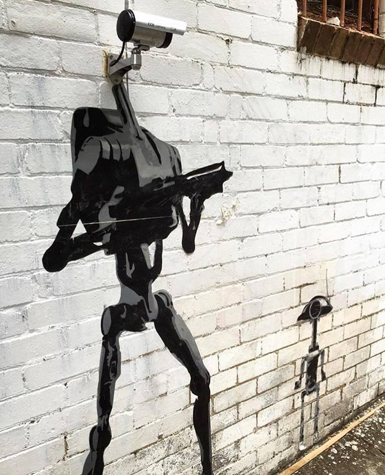 “CCTV Droid” street art by Murdoc in Sydney, Australia, using a real CCTV camera as the droid’s head above a black painted body on a white brick wall.