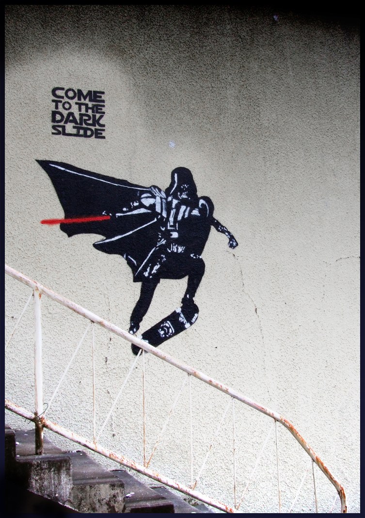 “Come To The Dark Slide” street art by Blouh, showing Darth Vader skateboarding on a wall above a stair railing while holding a red lightsaber.