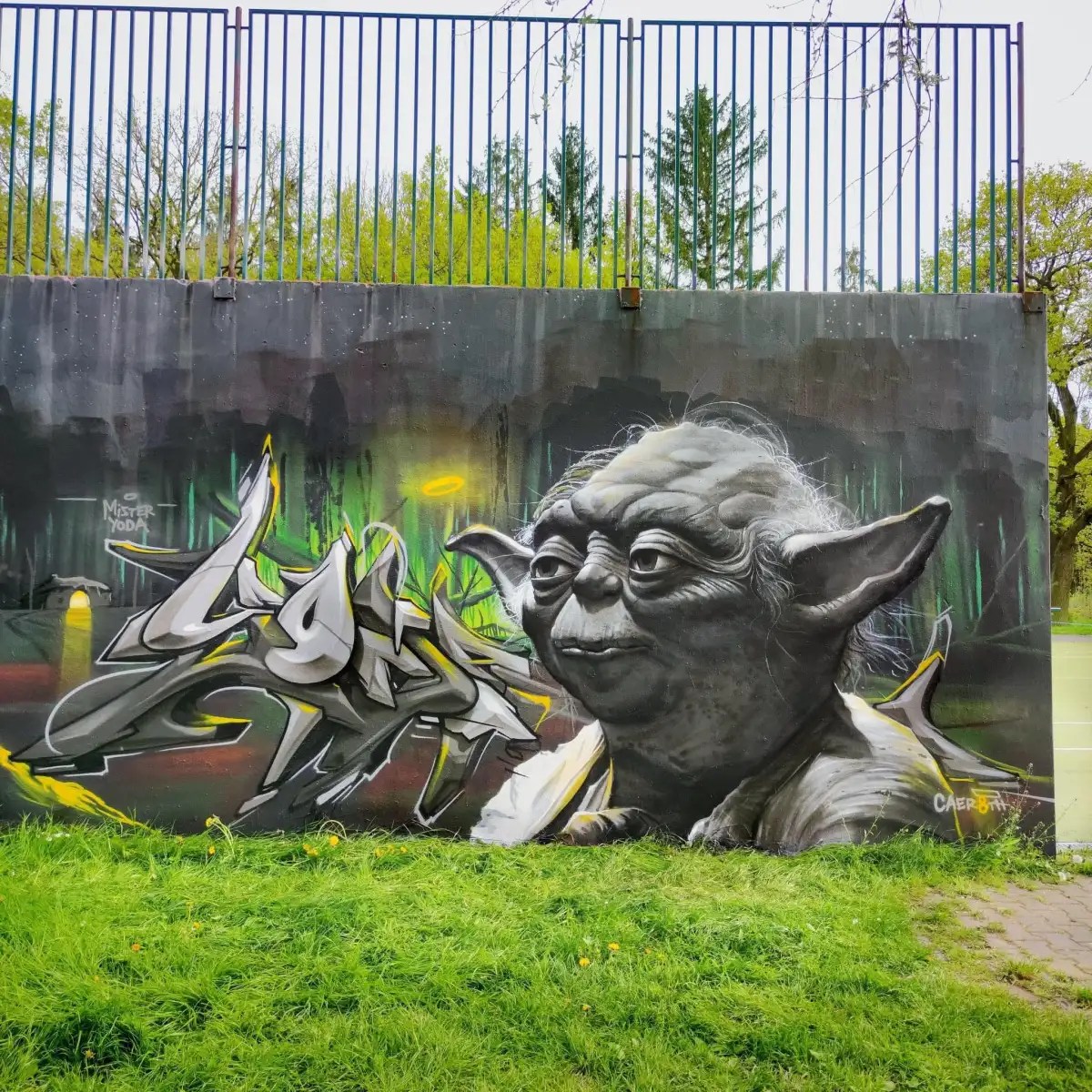 Gray-toned Yoda mural by Caer8th at Prague Castle, Czech Republic, with graffiti lettering on a wall below a metal fence.