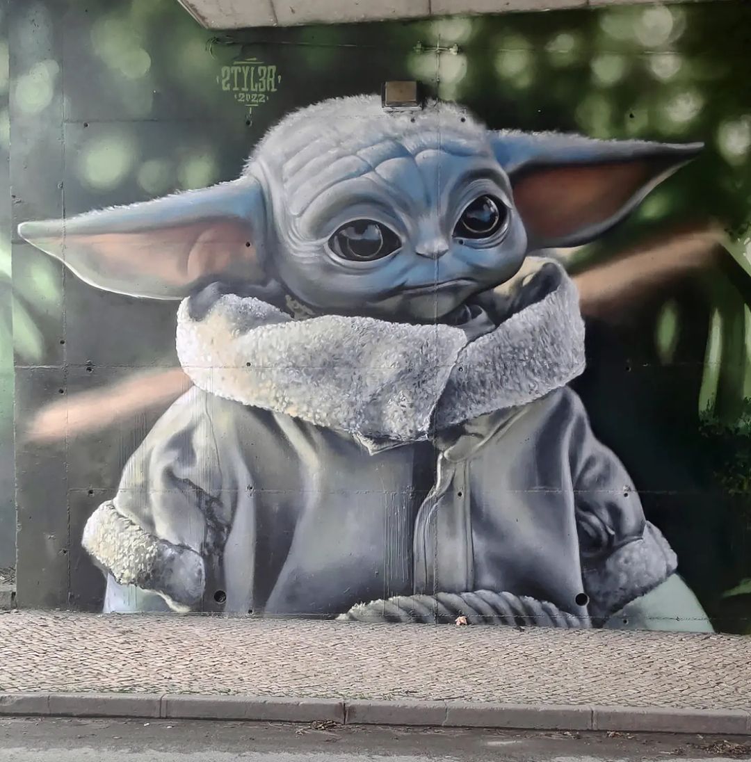 Grogu or Baby Yoda mural by Styler at the Viaduto da Ramada in Odivelas, Portugal, painted in a gray robe on a wall under a concrete overhang. Photo by Taemek.