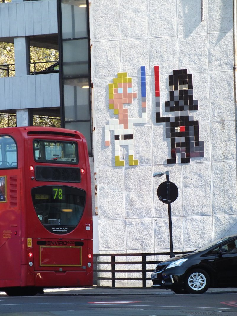 Invader’s LDN_132 Star Wars tile mosaic in London, England, showing pixelated Luke Skywalker and Darth Vader with blue and red lightsabers on a wall beside a red bus.