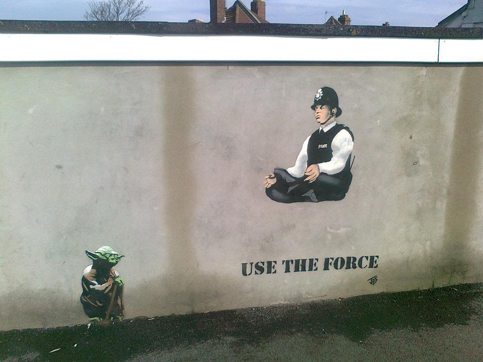 “Use The Force” street art by JPS, showing a small Yoda figure on the lower left and a seated police officer floating above the words on a plain wall.