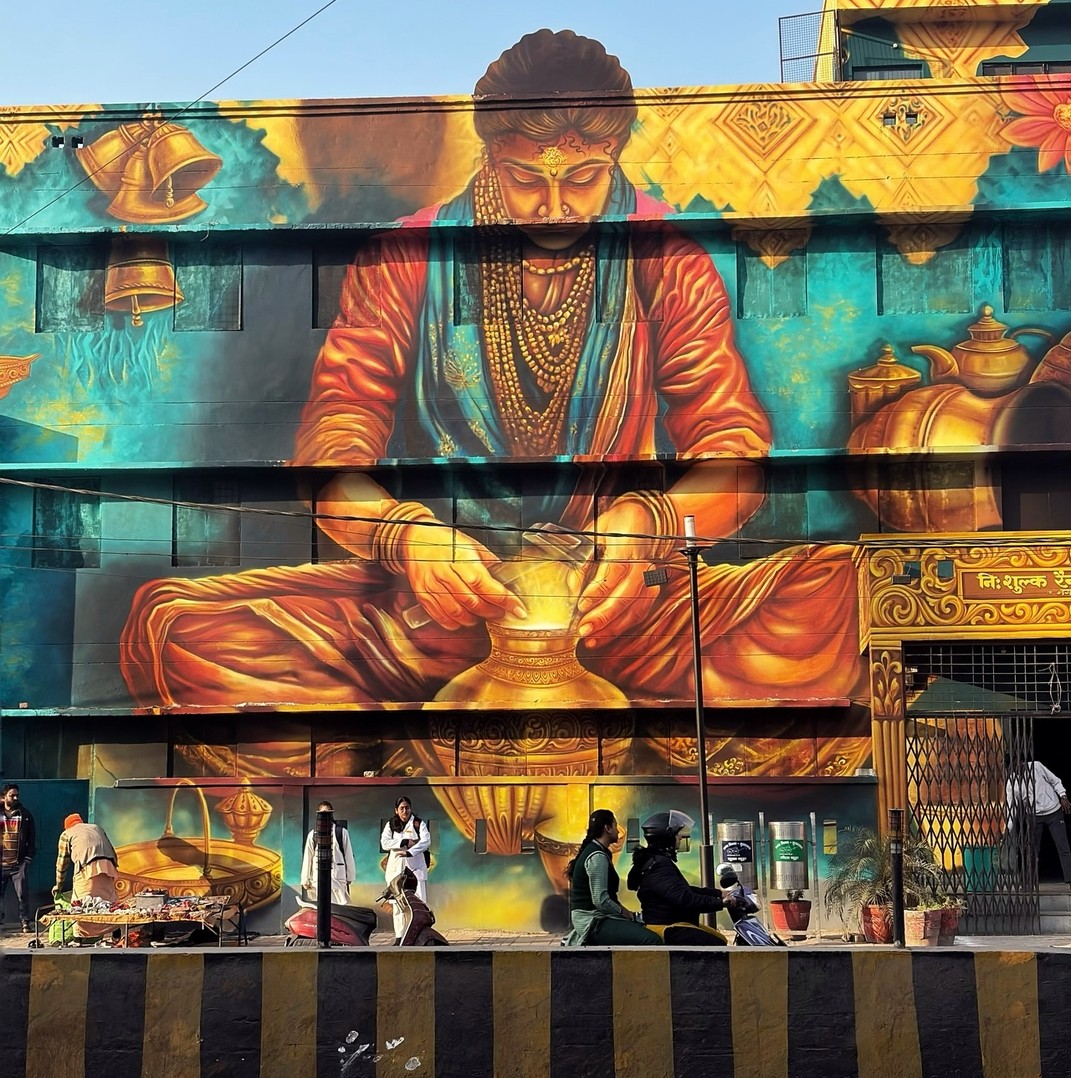 Alt text: A massive mural of a regal figure in traditional Indian attire, adorned with golden jewelry, pouring water from a vessel. The background is decorated with intricate patterns, bells, and golden vessels, adding a rich cultural feel. The mural integrates seamlessly with the building, with real-life street activity in front.