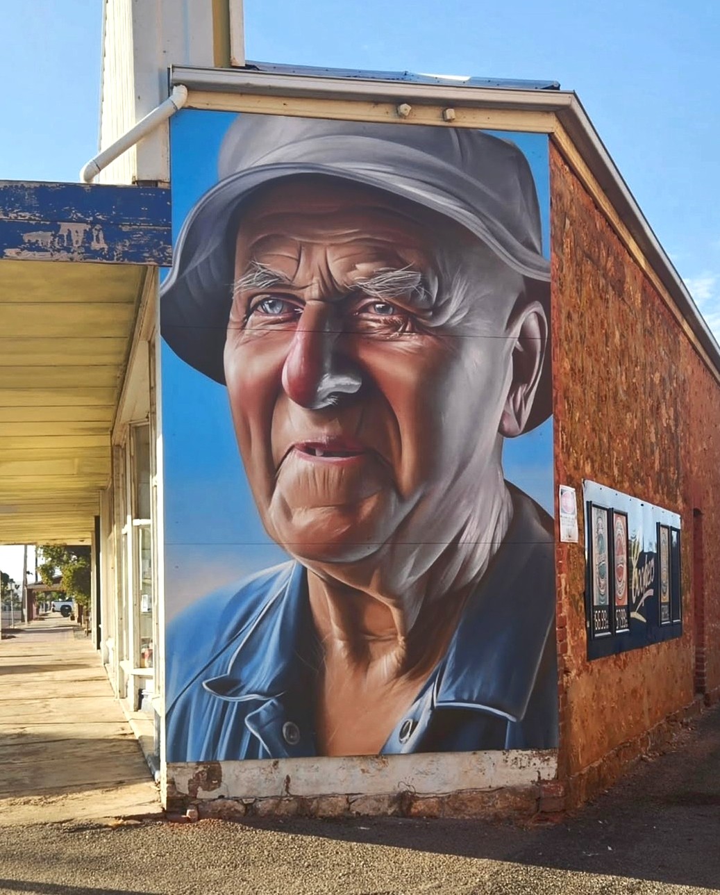 A highly detailed portrait of an elderly man wearing a bucket hat and blue work shirt. The wrinkles, sun-kissed skin, and reflective eyes are rendered in a hyper-realistic style, making the mural look almost like a photograph.