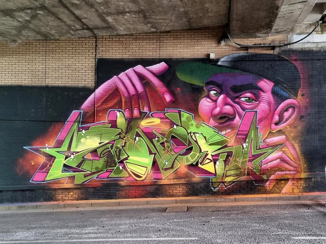 A vibrant graffiti mural featuring a stylized man with a serious expression, wearing a cap, surrounded by intricate lettering in green and purple. The mural has a bold urban aesthetic with dynamic lighting effects.