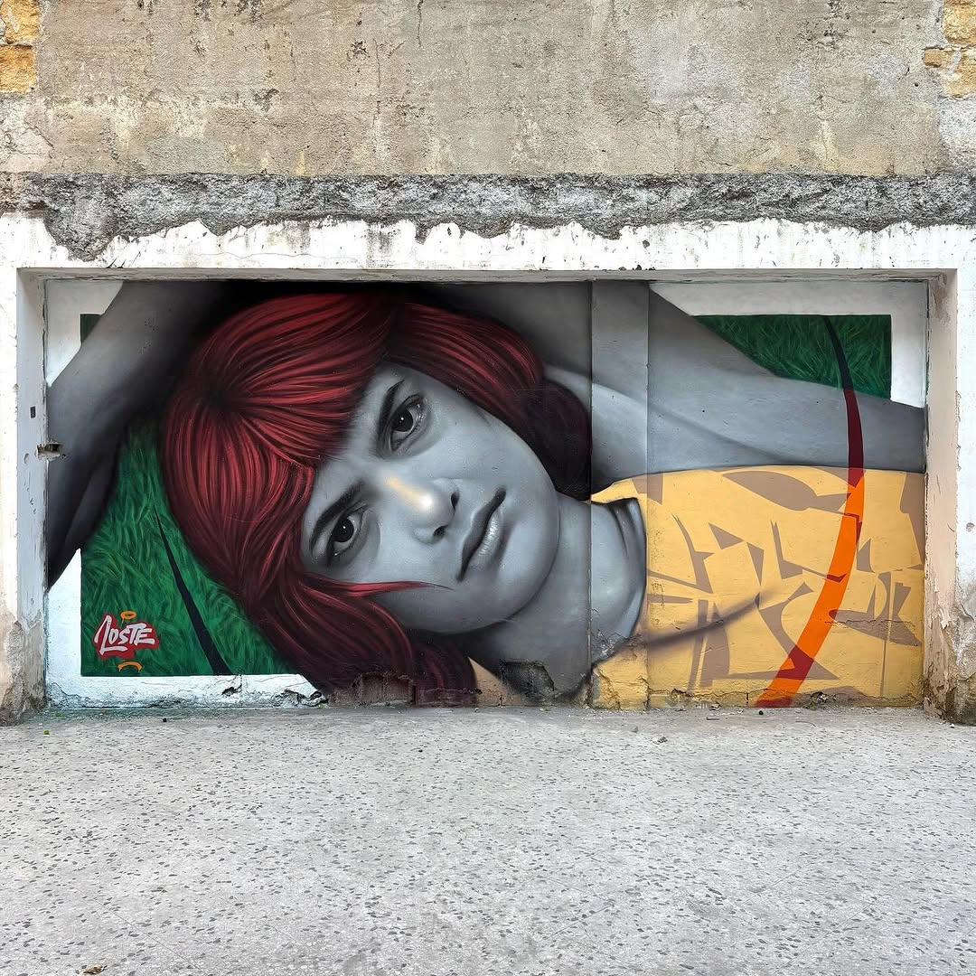 A mural of a woman with vivid red hair, painted in grayscale, lying in a resting pose with a distant gaze. Geometric shapes and abstract details in yellow and green surround her, creating a surreal atmosphere.