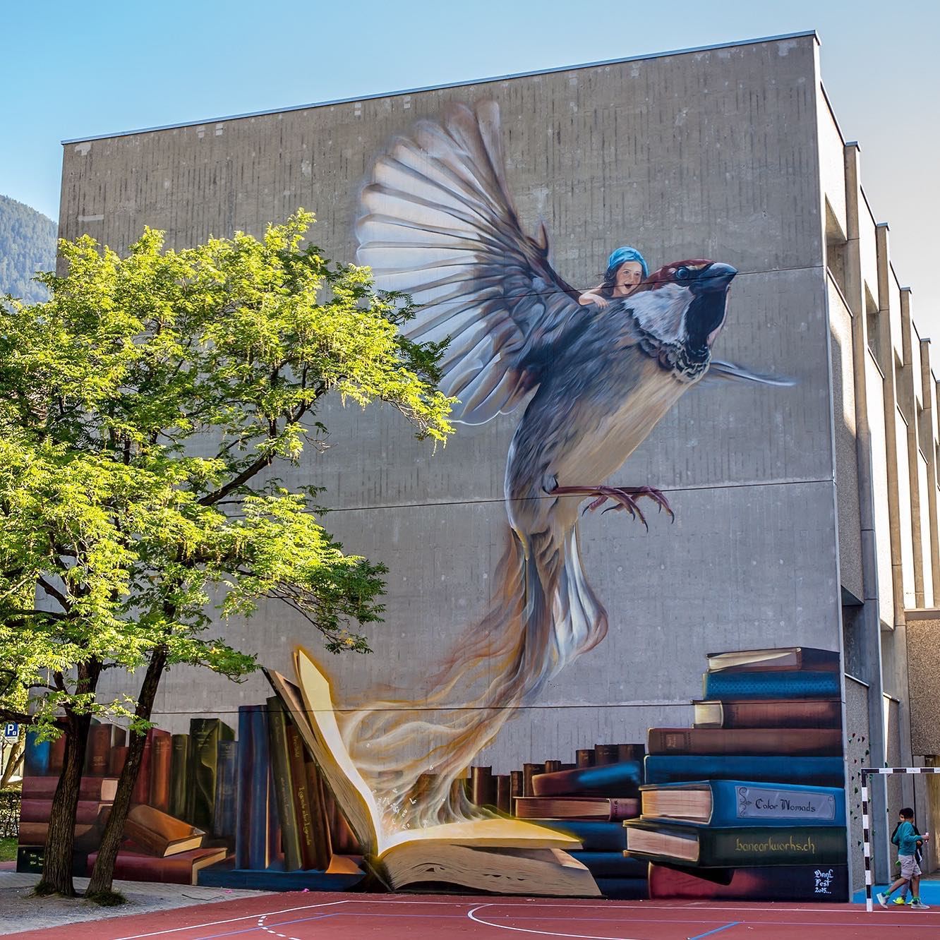 Large-scale mural of a girl riding a flying sparrow that rises from an open book surrounded by colorful stacks of books, painted on a school building.