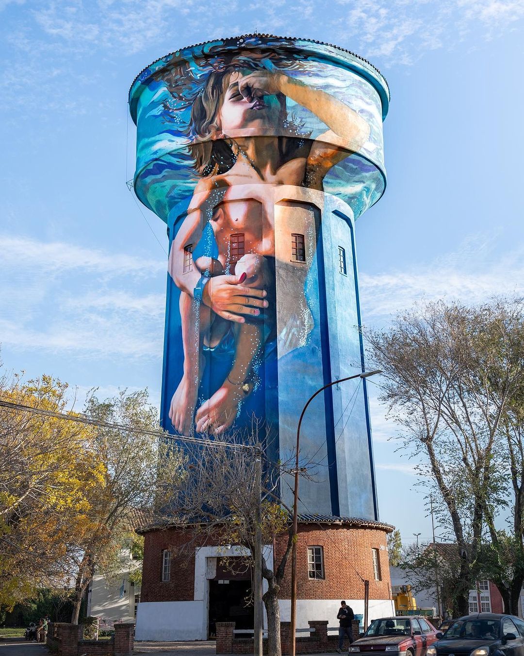 Photorealistic mural by Martin Ron on a water tower in Miramar, Argentina, showing a child underwater with eyes closed, nose pinched, and legs pulled up in a floating position. The painted waterline curves around the top of the tower, enhancing the depth of the scene.