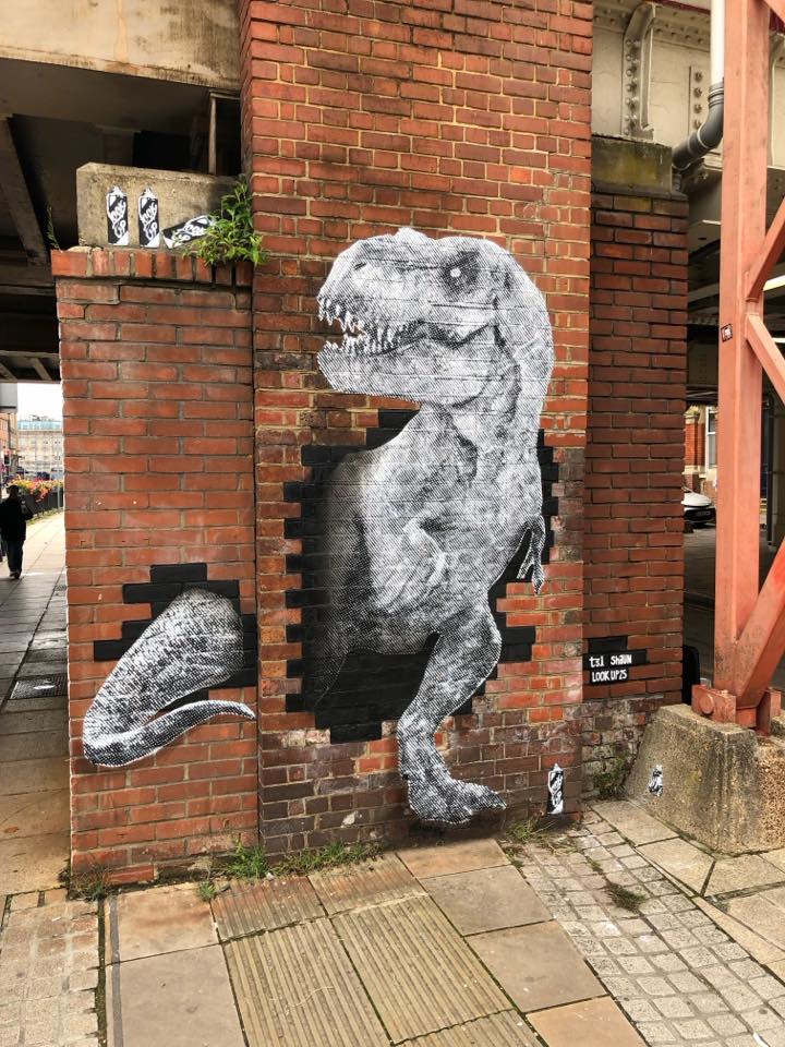 Black and white 3D stencil mural of a T-Rex breaking through a brick wall by Shaun Hodgkin.