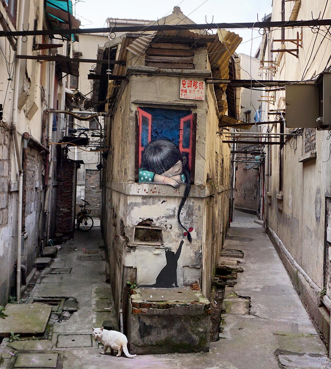 Street art by Seth Globepainter in Shanghai, China, featuring a girl with eyes closed leaning out of a painted red window. Her hair trails down the wall, ending in a flower being batted by a black painted cat. A real white cat walks below.