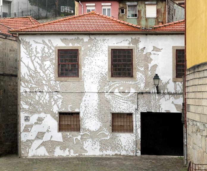 Vhils mural in Porto carved into a building facade, blending a human face with branch-like textures.