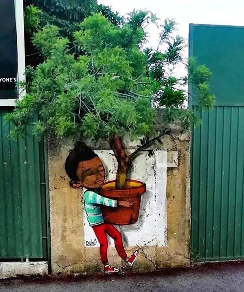Mural of a child hugging a painted red flowerpot around a real tree growing above a concrete wall, with green metal gates on both sides.