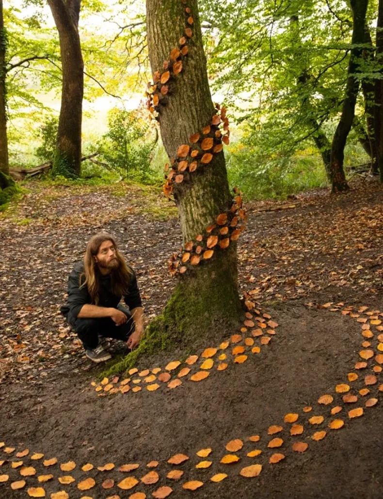 Vortex at Little Milford Woods by Jon Foreman, with orange autumn leaves arranged in a spiral around a tree trunk and across the forest floor while the artist crouches nearby.