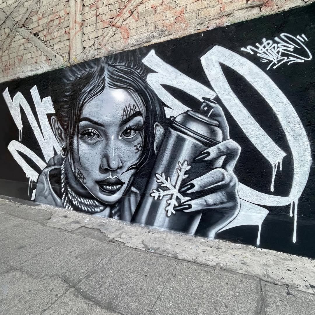 Black-and-white mural of a woman with intense gaze holding a spray can marked with a snowflake, detailed face tattoos, and large hoop earrings, with white graffiti lettering surrounding her, painted on a rough brick and stucco wall in Mexico City.