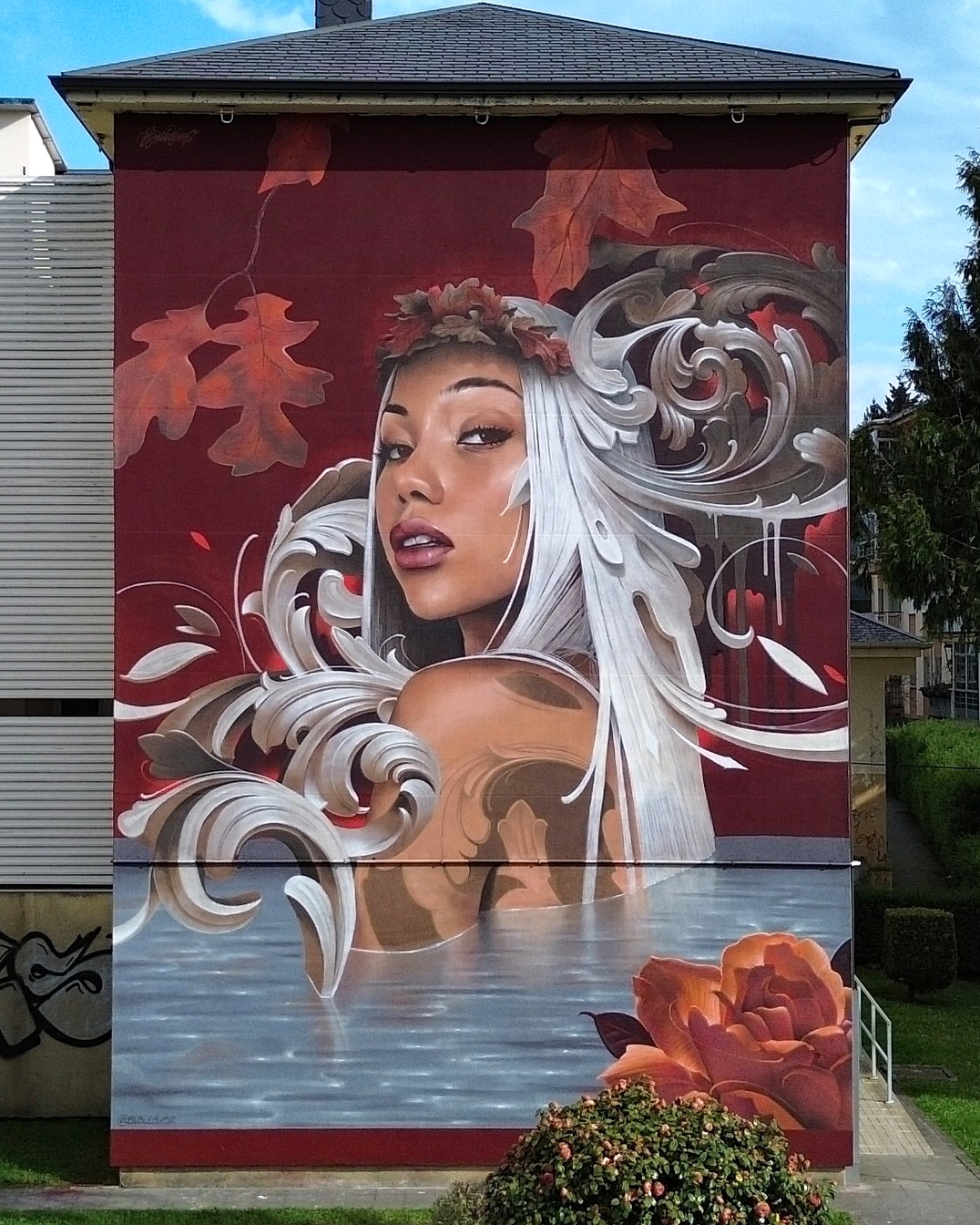 Mural of a silver-haired woman with elongated ears and autumn oak leaves swirling around her head and body, partially submerged in water with stylized swirls, painted on a burgundy wall in Lugo, Spain.