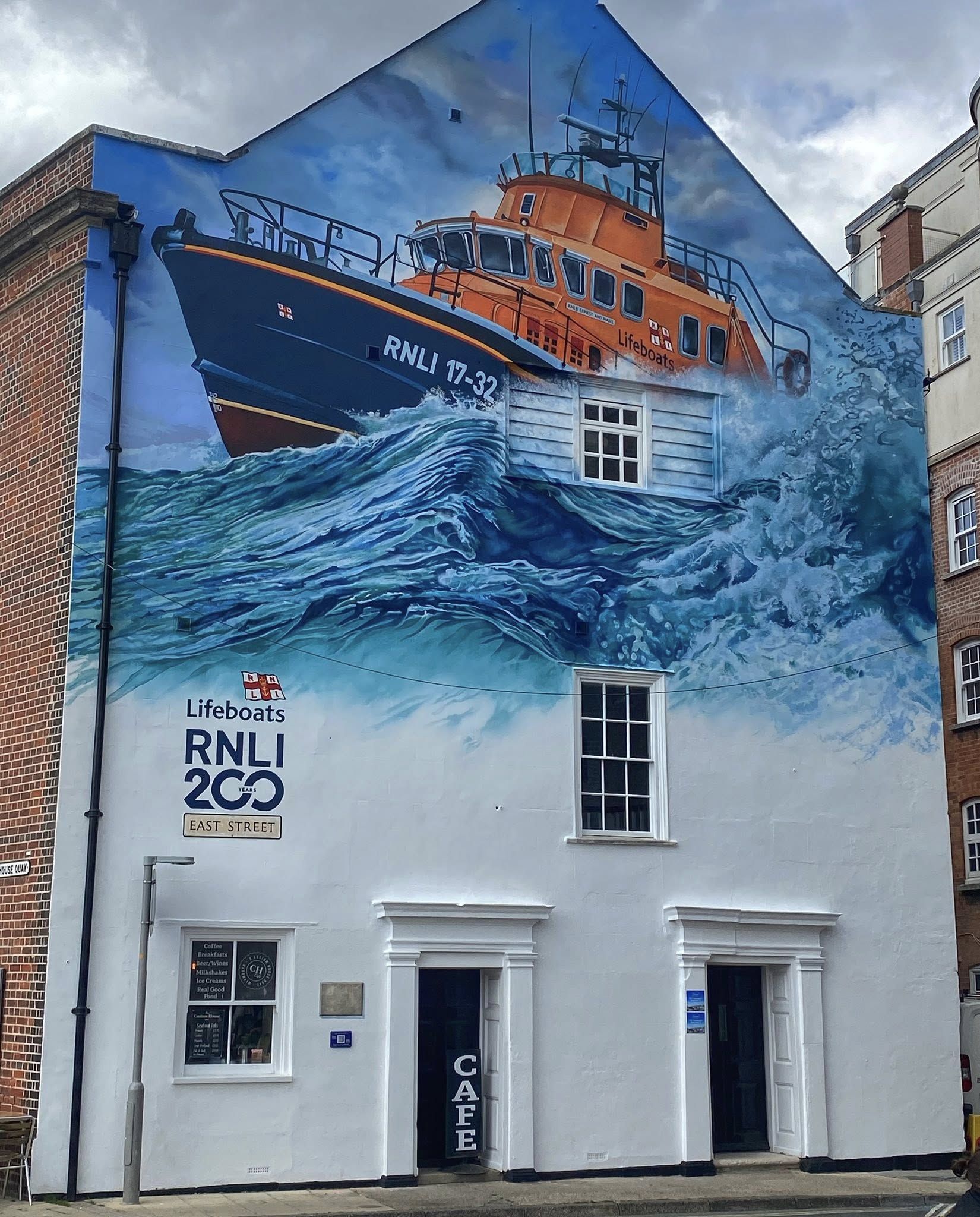 Realistic mural of an RNLI orange lifeboat bursting through ocean waves on the side of a white historic building with real windows and doors, commemorating 200 years of the RNLI in Weymouth, England.