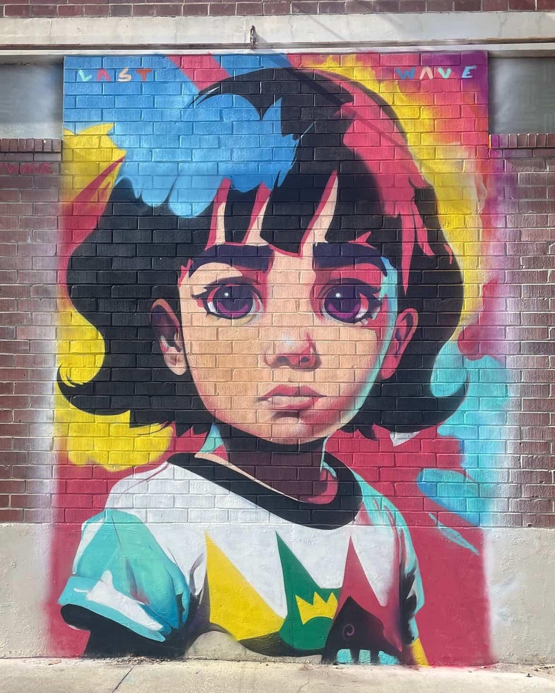 Colorful mural of a young girl with solemn expression and large violet eyes, wearing a graphic t-shirt, painted in pink, blue, and yellow tones against a red brick wall in Jacksonville, Florida.