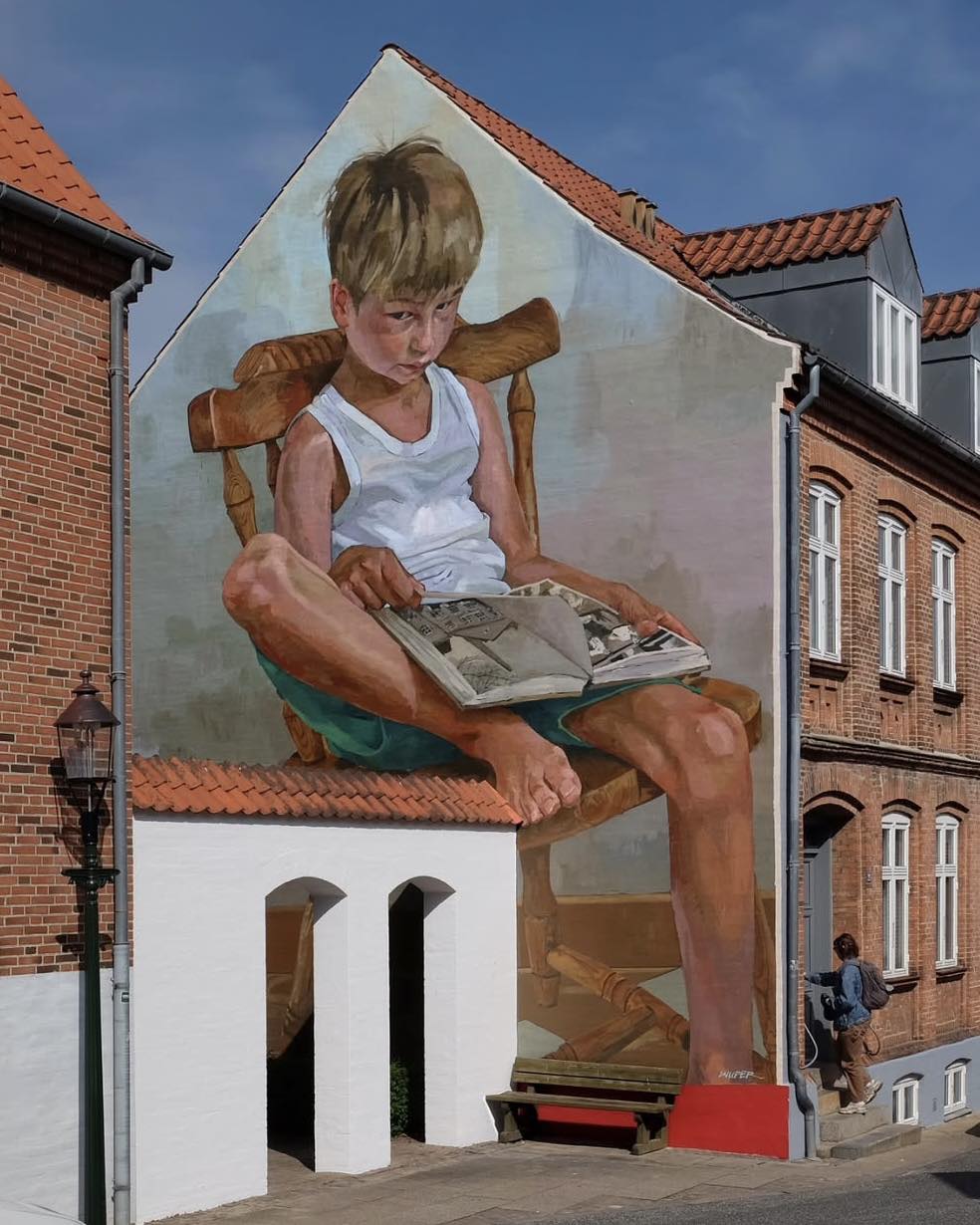 Large mural of a blond boy sitting barefoot on a massive wooden chair, absorbed in a book, painted to fit the contours and roof of a residential building in Viborg, Denmark.
