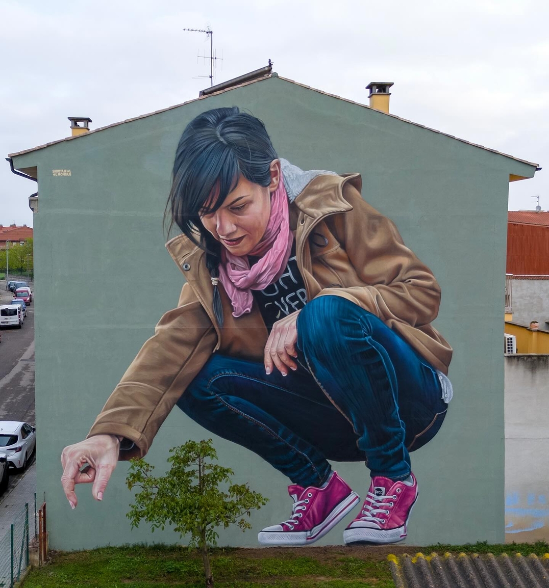 Photorealistic mural of a woman crouching down with her hand extended toward a real tree growing in front of the wall, painted on a green building in Civitacampomarano, Italy. She wears a brown coat, blue jeans, pink scarf, and pink sneakers, appearing lifelike and integrated into the urban landscape.