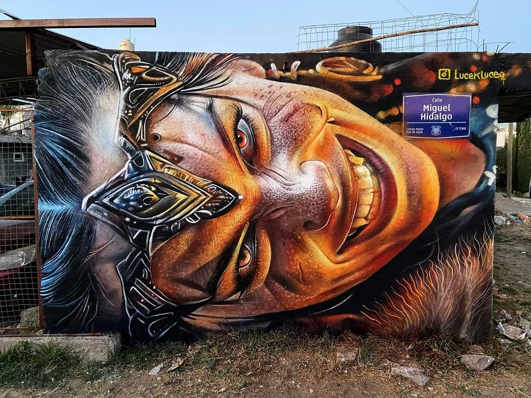 Detailed street art portrait of a smiling person with golden teeth and a metallic headpiece, painted with warm orange and red tones on a wall in Puebla, Mexico.