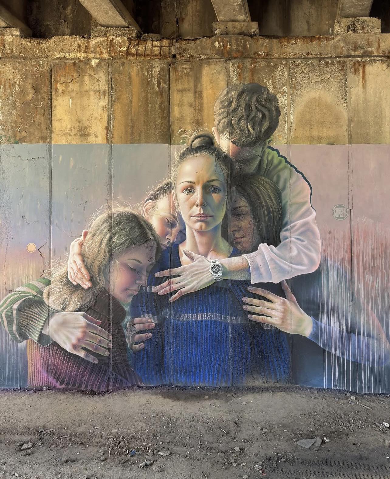 Mural showing a group of five people embracing closely, painted with soft colors and realistic shading on a rough concrete wall under a bridge in Kyiv, Ukraine.