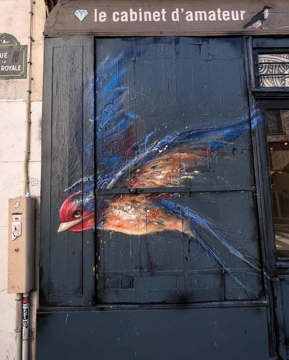 Mural on Paris storefront depicting one dynamic, painterly bird in flight against a deep blue background, with visible motion lines and vibrant feathers.