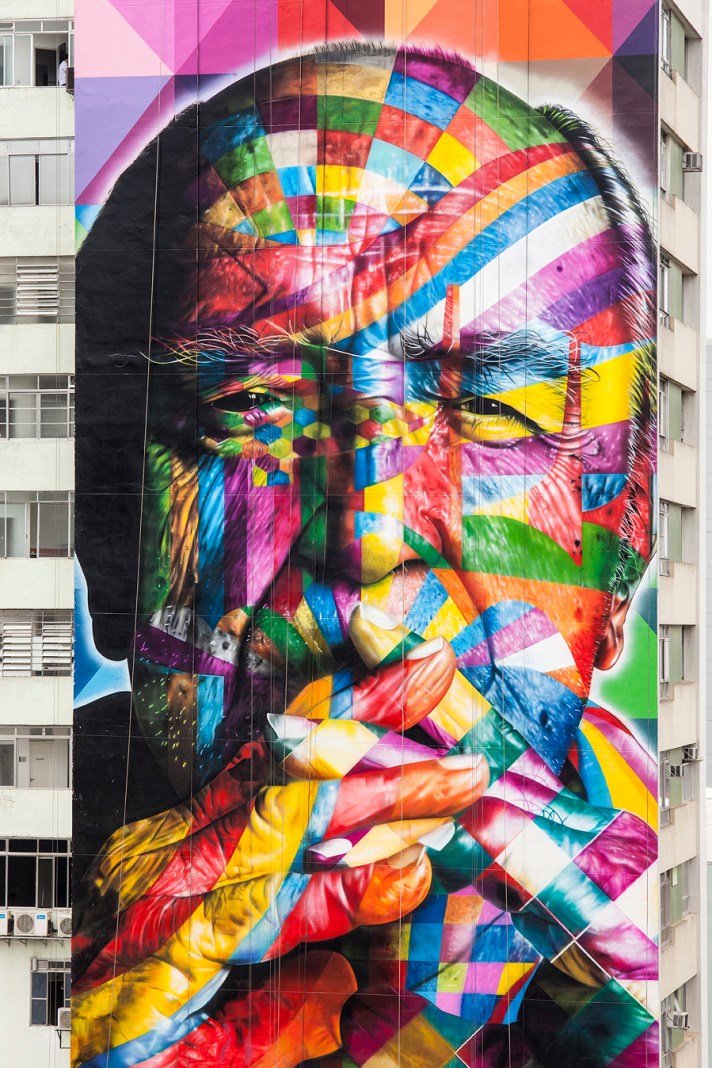 A monumental mural by the artist Kobra in São Paulo, Brazil, paying tribute to the renowned architect Oscar Niemeyer. The artwork features a colorful, geometric portrait of Niemeyer, with intricate patterns and bold lines forming a mosaic-like effect across his face and hand. The design is vibrant, with shades of red, blue, yellow, green, and purple blending seamlessly. Niemeyer is depicted in a thoughtful pose, holding a cigarette, symbolizing his creative and reflective nature. The mural spans the side of a tall building, standing as a dynamic tribute to his architectural legacy and artistic influence.