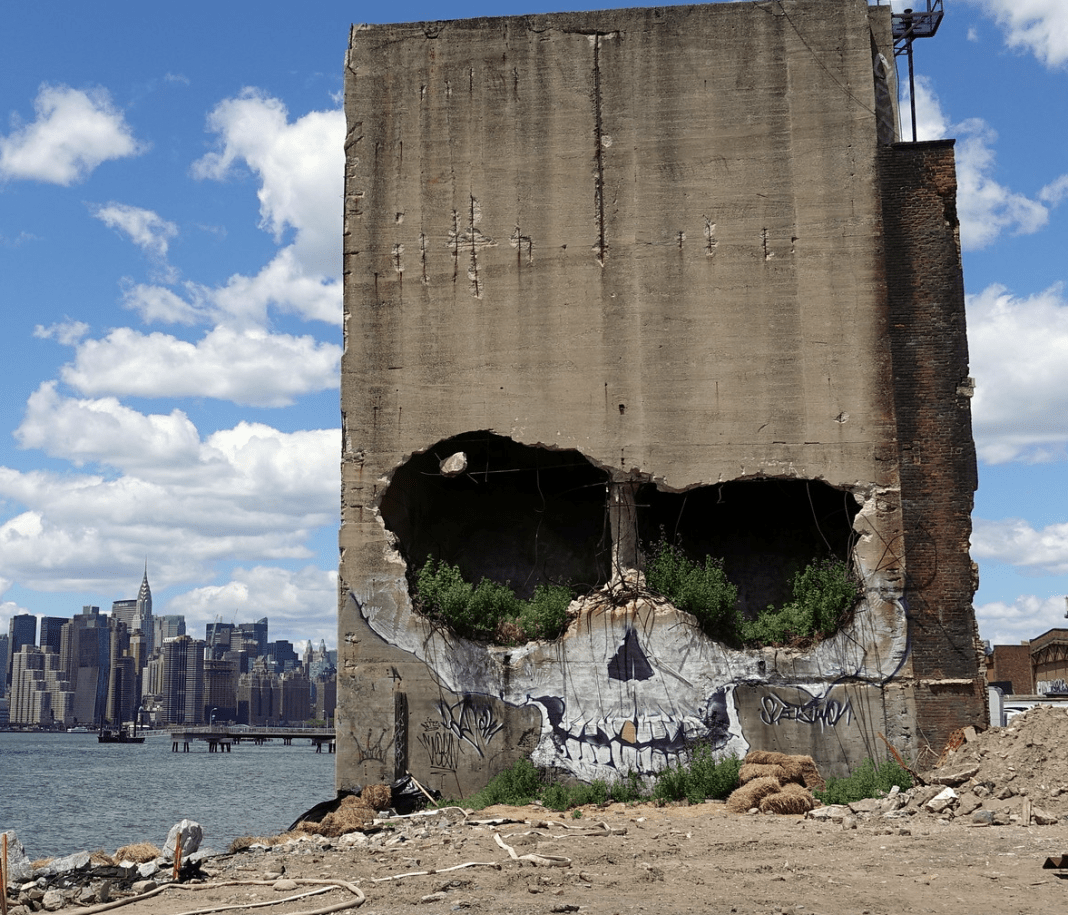 A haunting mural by Suitswon on a crumbling concrete structure in Brooklyn, New York, USA. The artwork transforms the dilapidated building into a giant skull, with two large holes resembling hollow eye sockets and cracks forming a menacing grin. Green plants grow within the openings, blending nature with urban decay. The Manhattan skyline is visible across the water under a bright blue sky, contrasting the eerie, skeletal facade. The rawness of the ruins and the surrounding debris amplify the gritty, post-apocalyptic aesthetic of the piece.