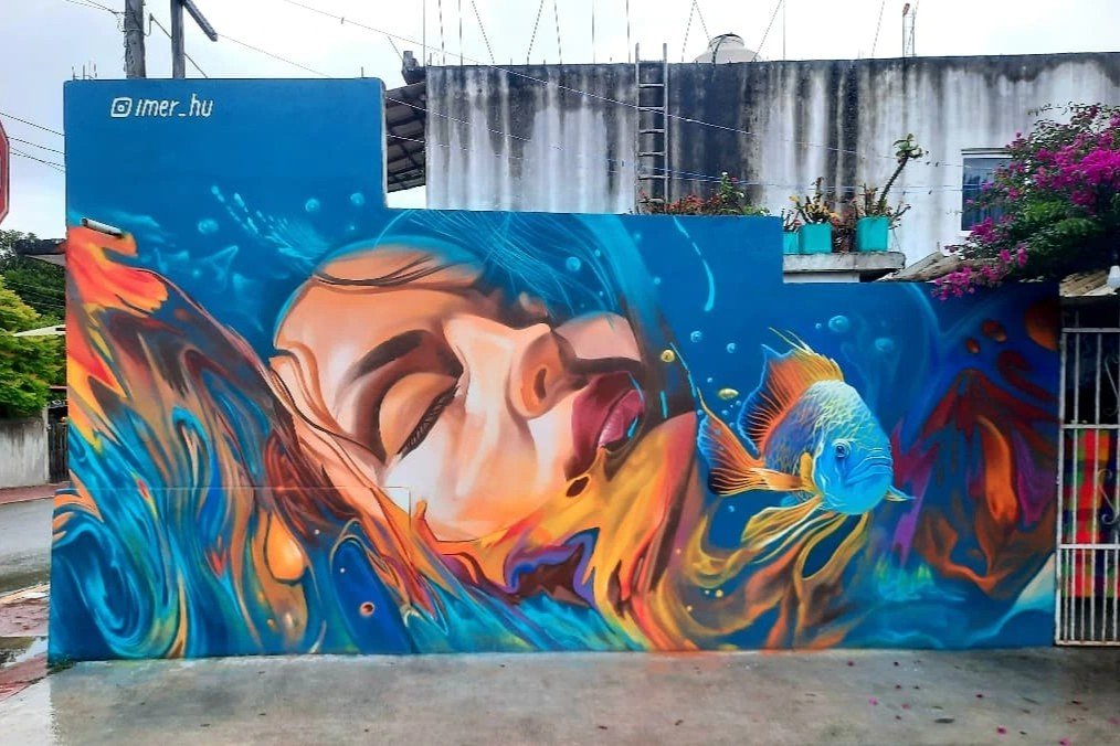 A mural by Imer Hu in Bacalar, Mexico, showcases a woman’s serene face partially submerged in swirling blue and orange water. A vibrant fish swims nearby, harmonizing with the fluid forms, creating an ethereal, dreamlike effect.