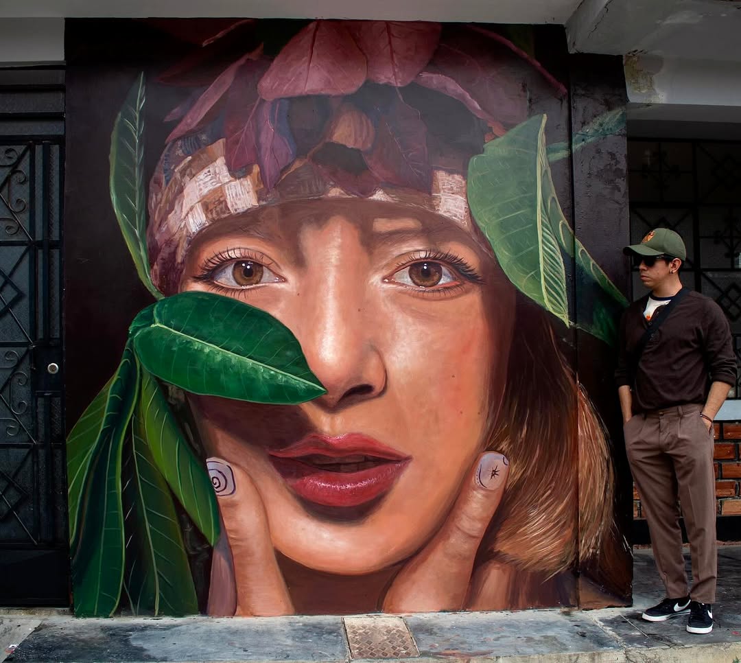 A mural by Machuca in Moyobamba, Peru, features a highly realistic portrait of a woman with deep brown eyes. Her head is adorned with red leaves, and green foliage partially covers her face, symbolizing a deep connection with nature.