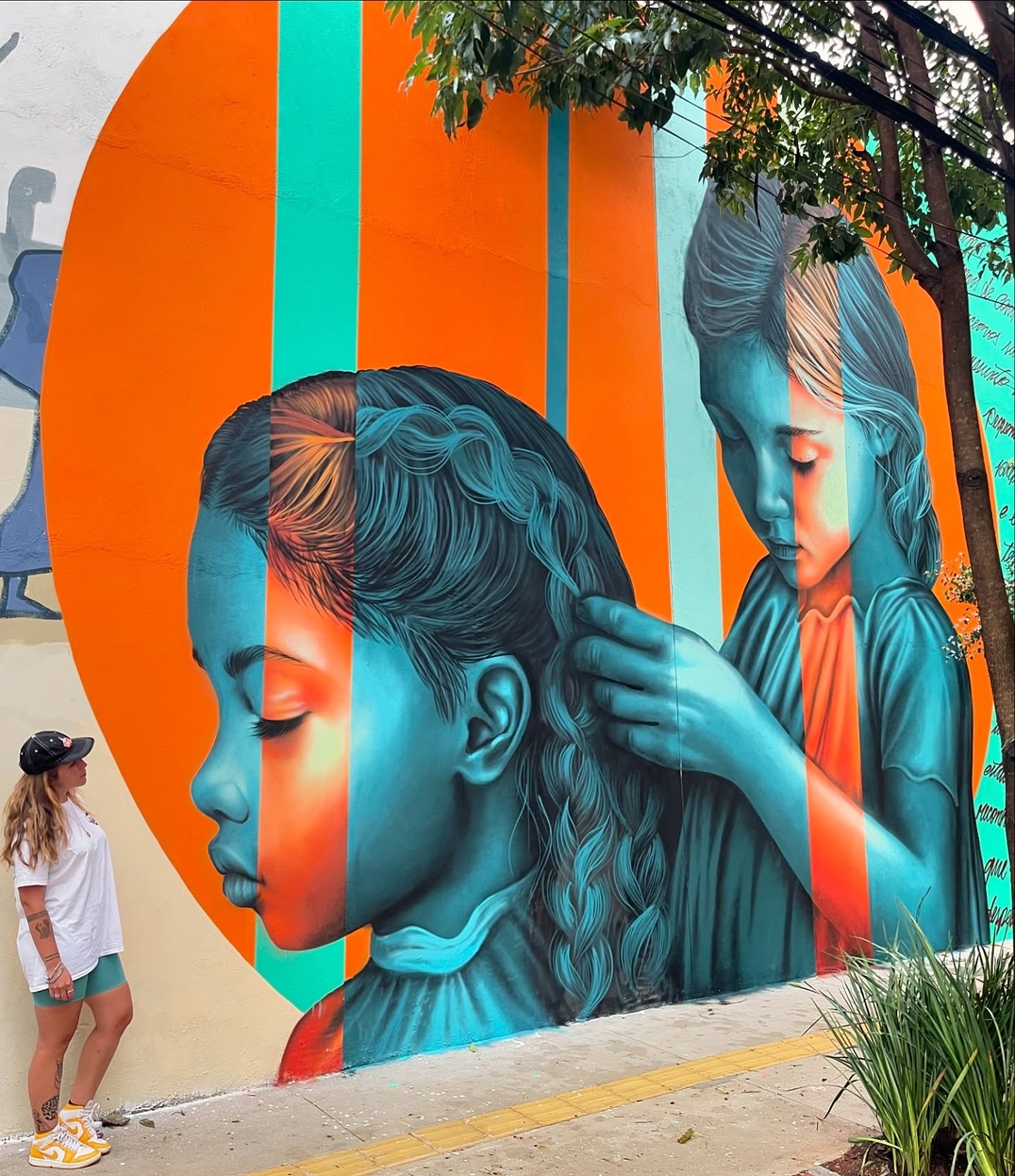 A mural by Clara Leff in São Paulo, Brazil, depicts two young girls in shades of blue, one gently braiding the other’s hair. The composition is framed by an orange circular backdrop, creating a contrast that emphasizes their bond.