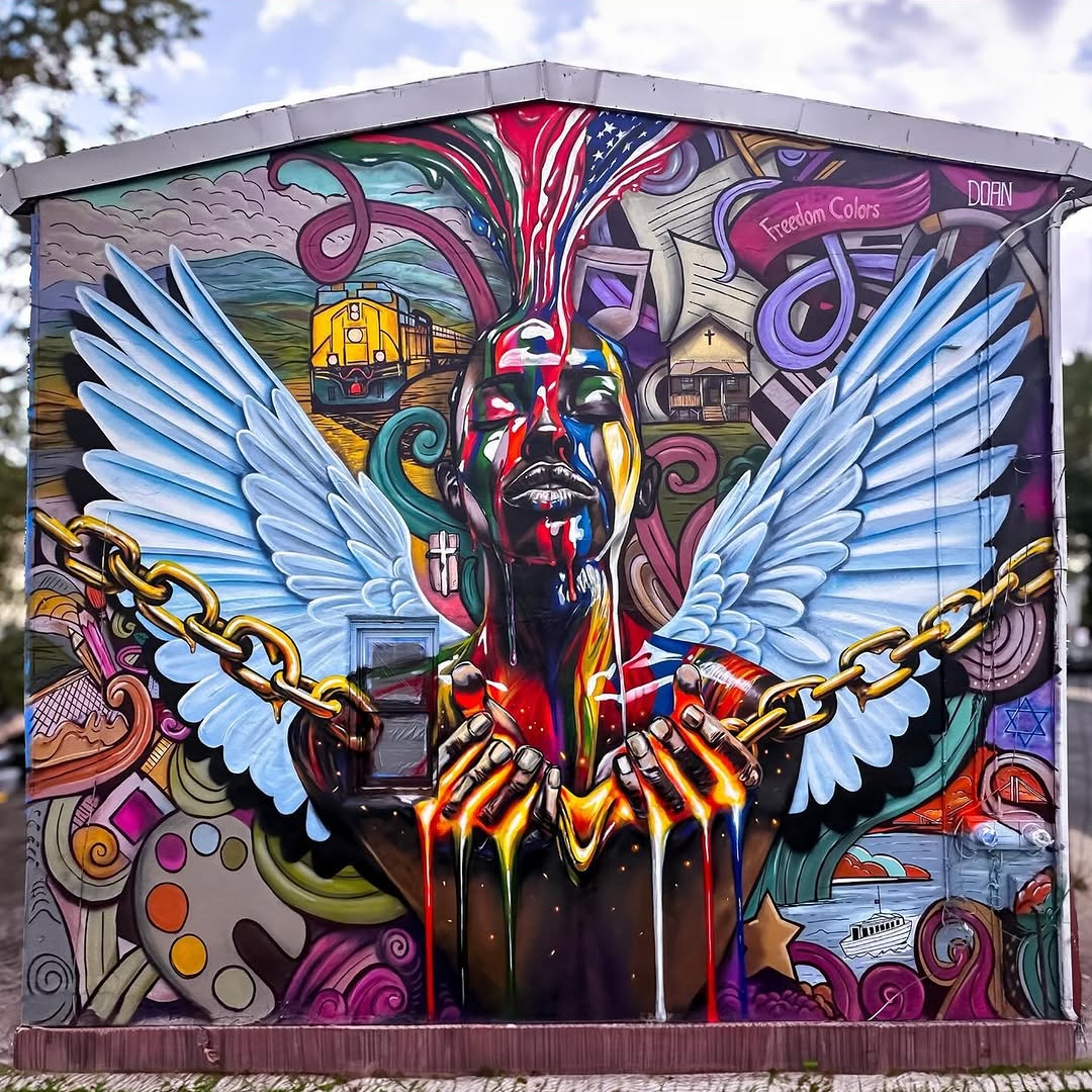 A mural by Sıtkı Doğan in Haverstraw, New York, USA, features a colorful angelic figure with large white wings and glowing skin. Chains break apart in front of them, symbolizing freedom. The background is filled with vibrant patterns, a train, and religious iconography.