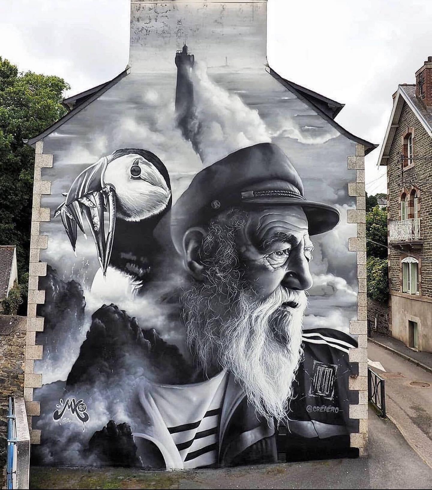 Black and white mural showing an old sea captain with a puffin and a lighthouse in the background, painted on a stone-walled house in France.