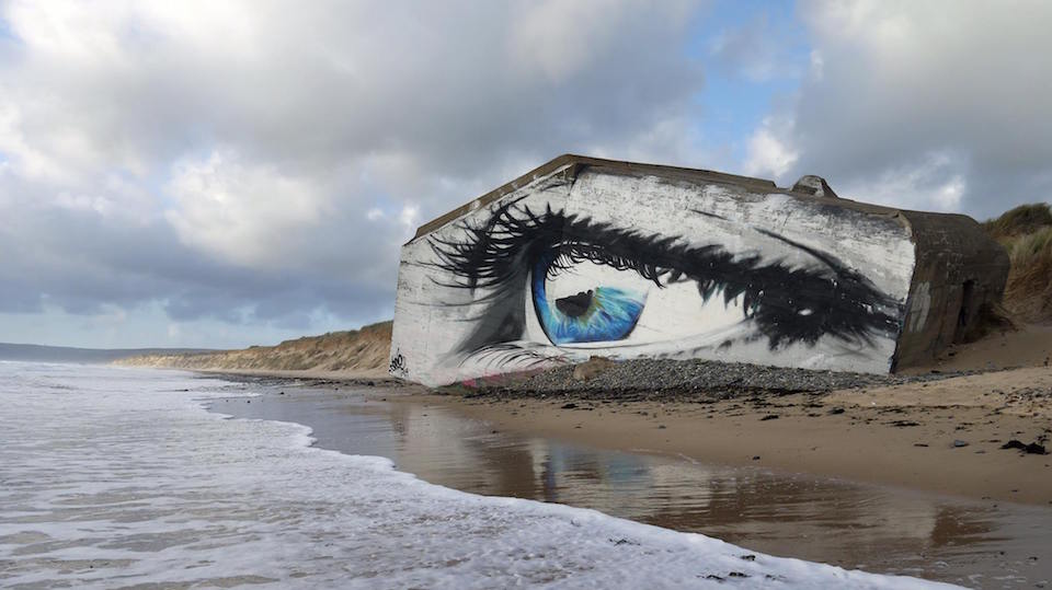 Mural by Näutil on a WWII bunker in Siouville-Hague, France, painted to resemble a realistic human eye with detailed eyelashes and a reflection of a ship in the blue iris, located directly on the beach with waves nearby.