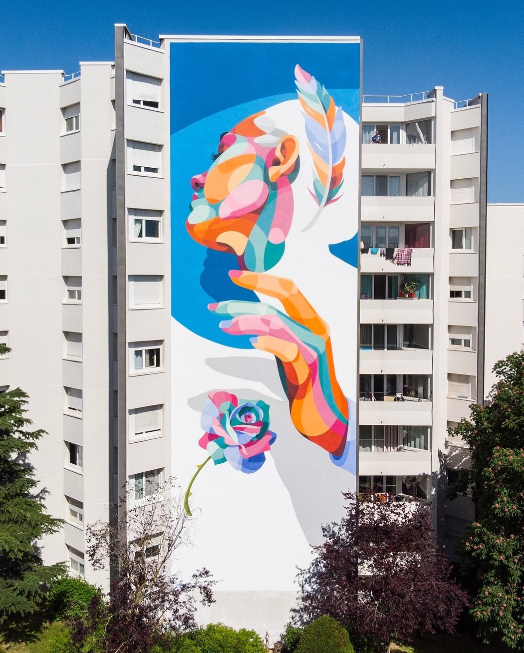 Mural by Alber in Saint-Germain-en-Laye, France, depicting a woman's profile in multicolored abstract segments, with her hand reaching toward a vibrant rose and a feather behind her head, painted on the full height of a residential building.