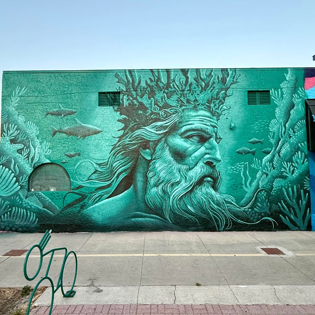 Mural by Derek Donnelly in Gulfport, Florida, showing an underwater god figure with a long beard, crowned in coral and surrounded by fish and aquatic plants in shades of green.