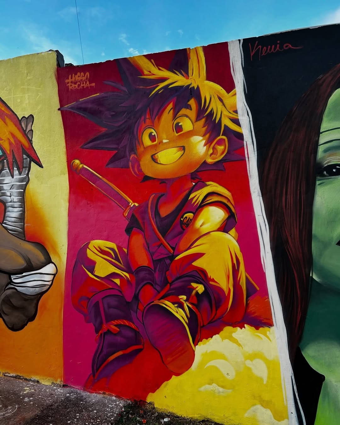 Mural by Huggo Rocha in Londrina, Brazil, featuring Goku in anime style with glowing yellow and red tones, sitting on a cloud with a sword on his back and a bright sky-blue background.